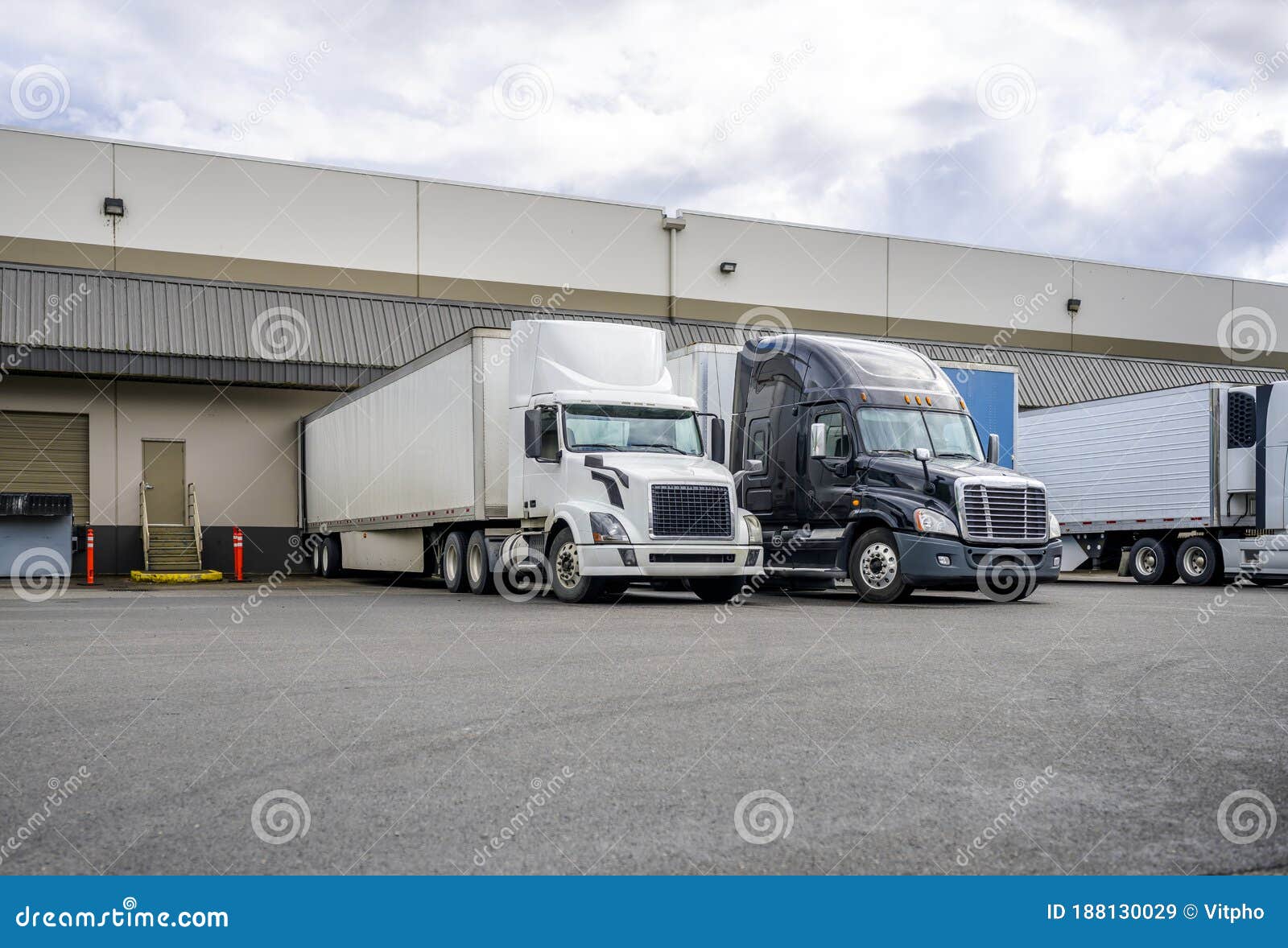 black and white big rigs semi trucks with semi trailers loading cargo at warehouse dock with gates for each