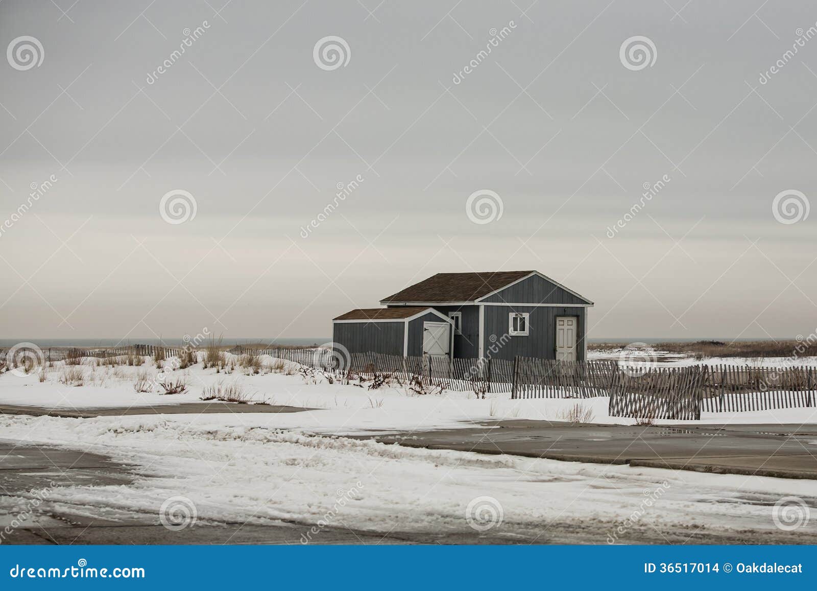 Blauw Strandhuis op Snow-covered Duin. Onder grijze, donkere hemel, is een klein blauw die strandhuis op een snow-covered duin wordt en door omheiningen dichtbij een parkeerterrein door het overzees wordt omringd gevestigd die omhoog gesloten voor de winter.