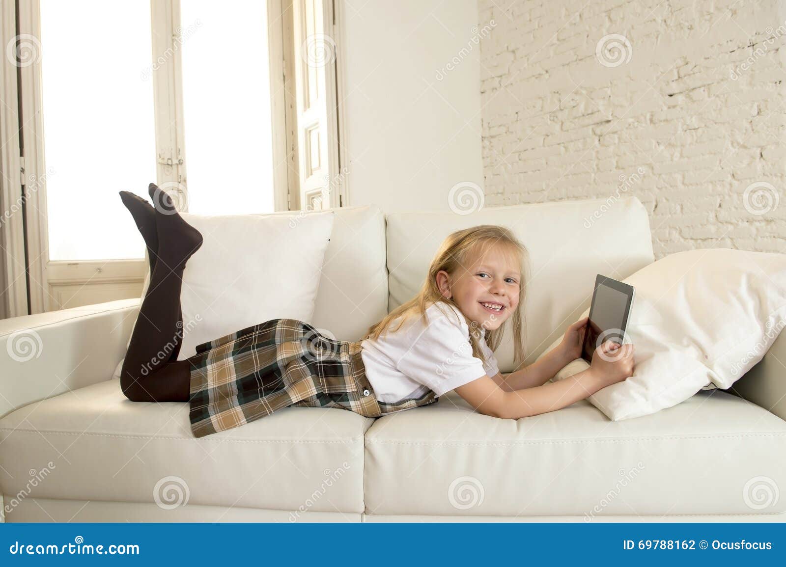 blond little girl lying on home sofa couch using internet app on digital tablet pad on digital tablet pad