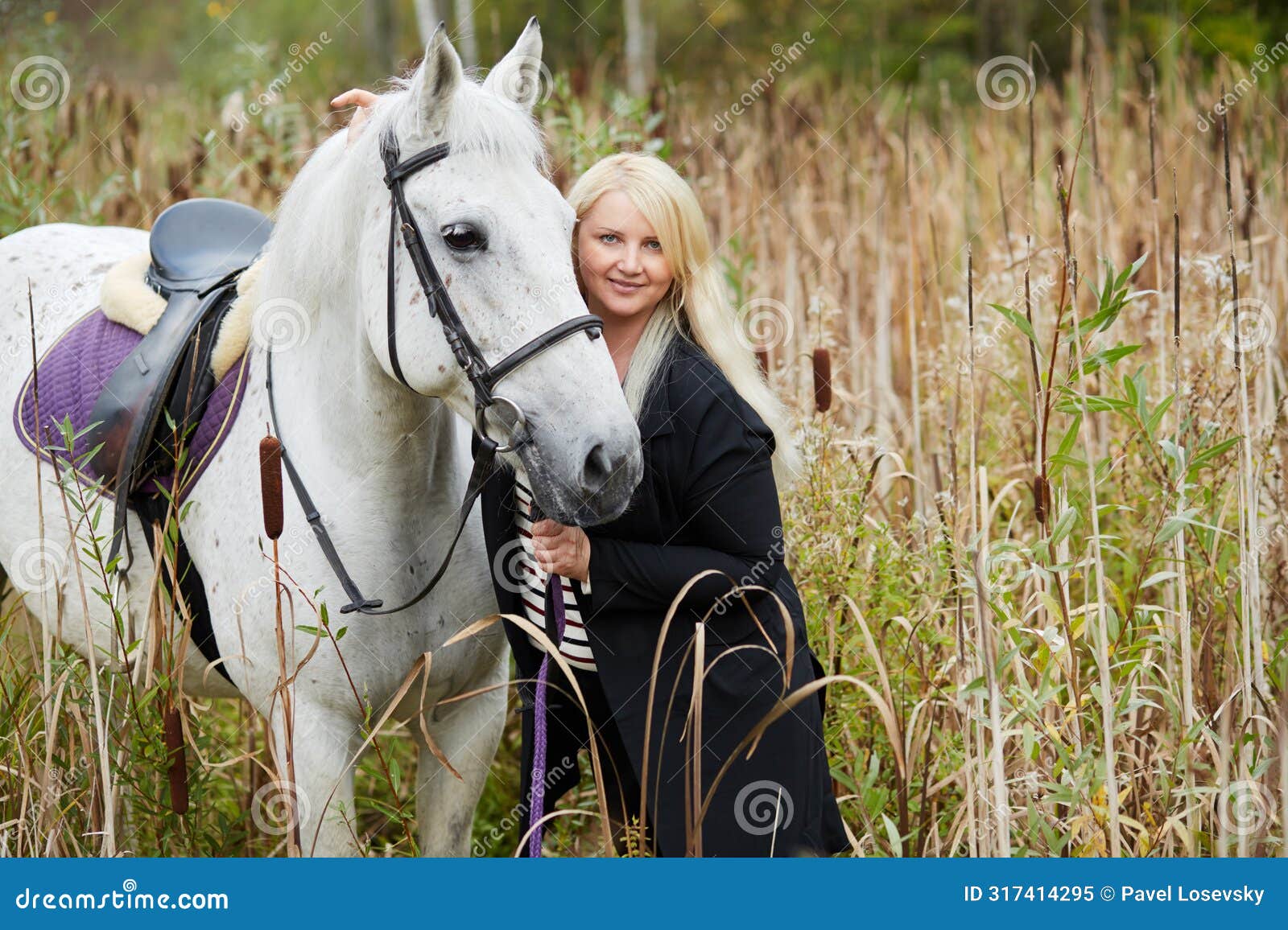 blonde woman holds by bridle white horse among the