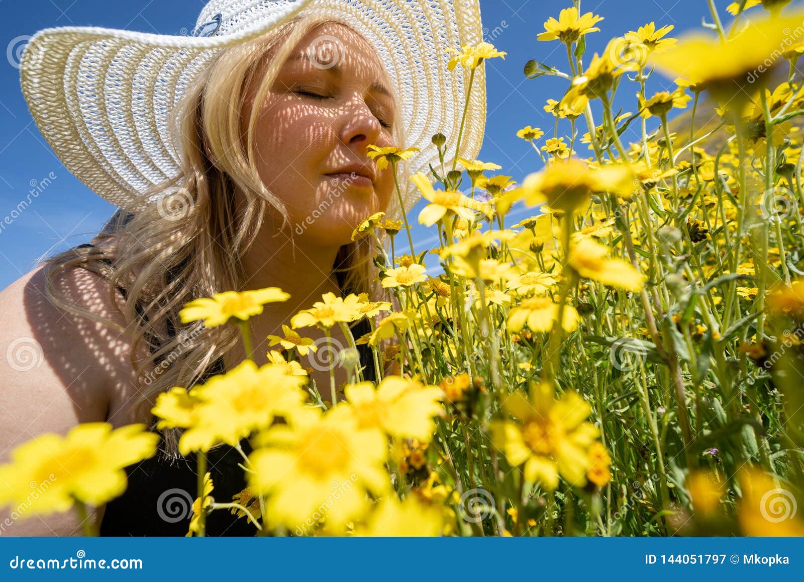 blonde woman wearing straw white hat smelling a field of yellow wildflowers. concept for springtime allergy relief