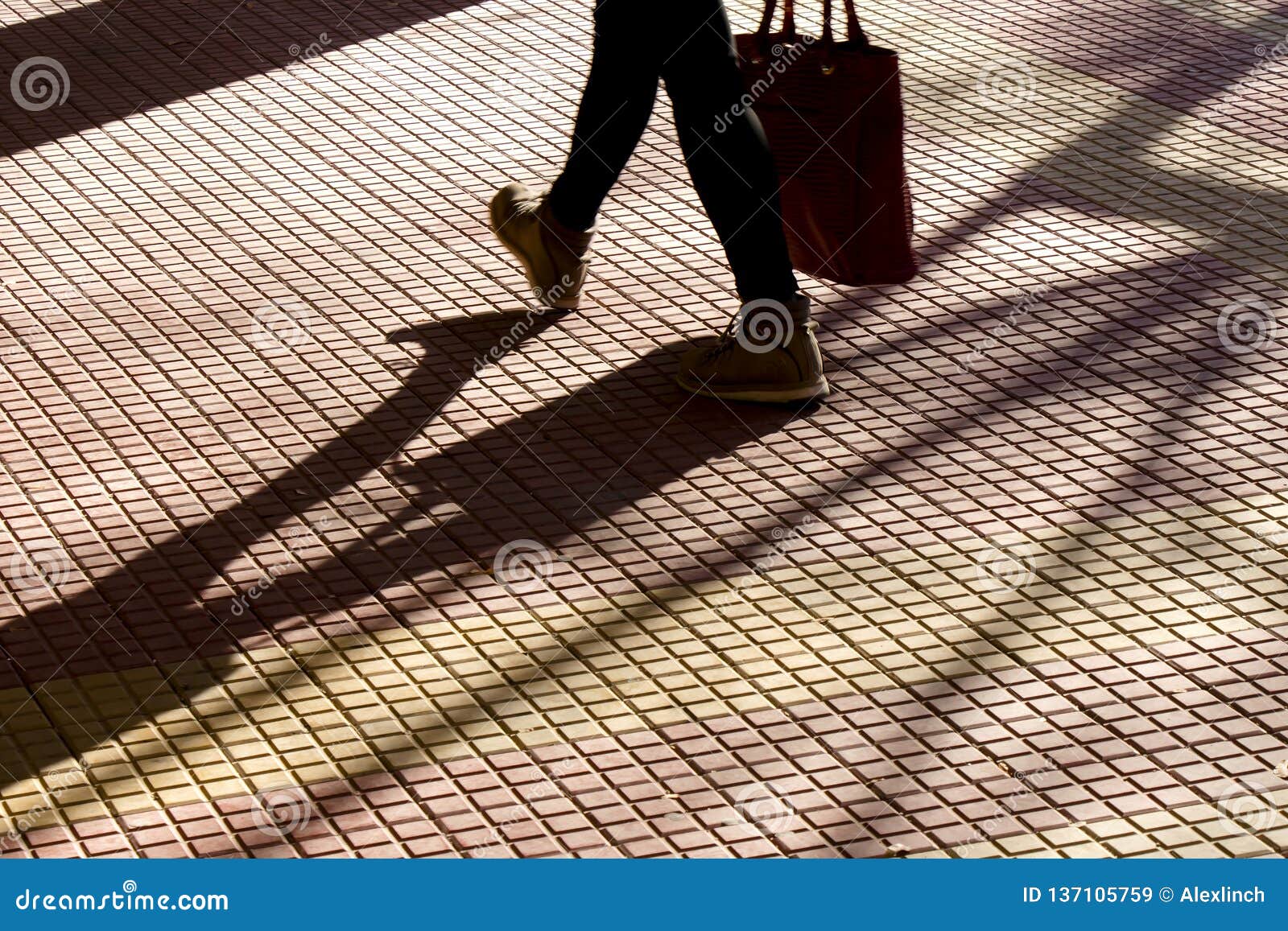 blurry silhouette shadow of legs of a person carrying a bag while walking on tiled street sidewalk