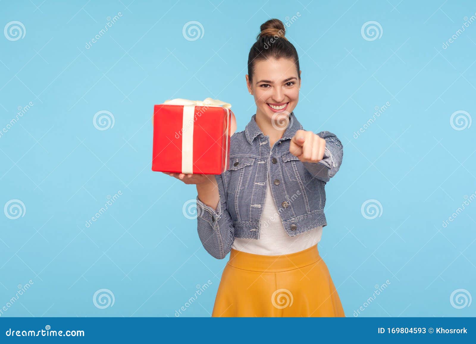 bonus for you, winner! portrait of optimistic beautiful fashionably dressed girl with hair bun holding gift box and pointing to