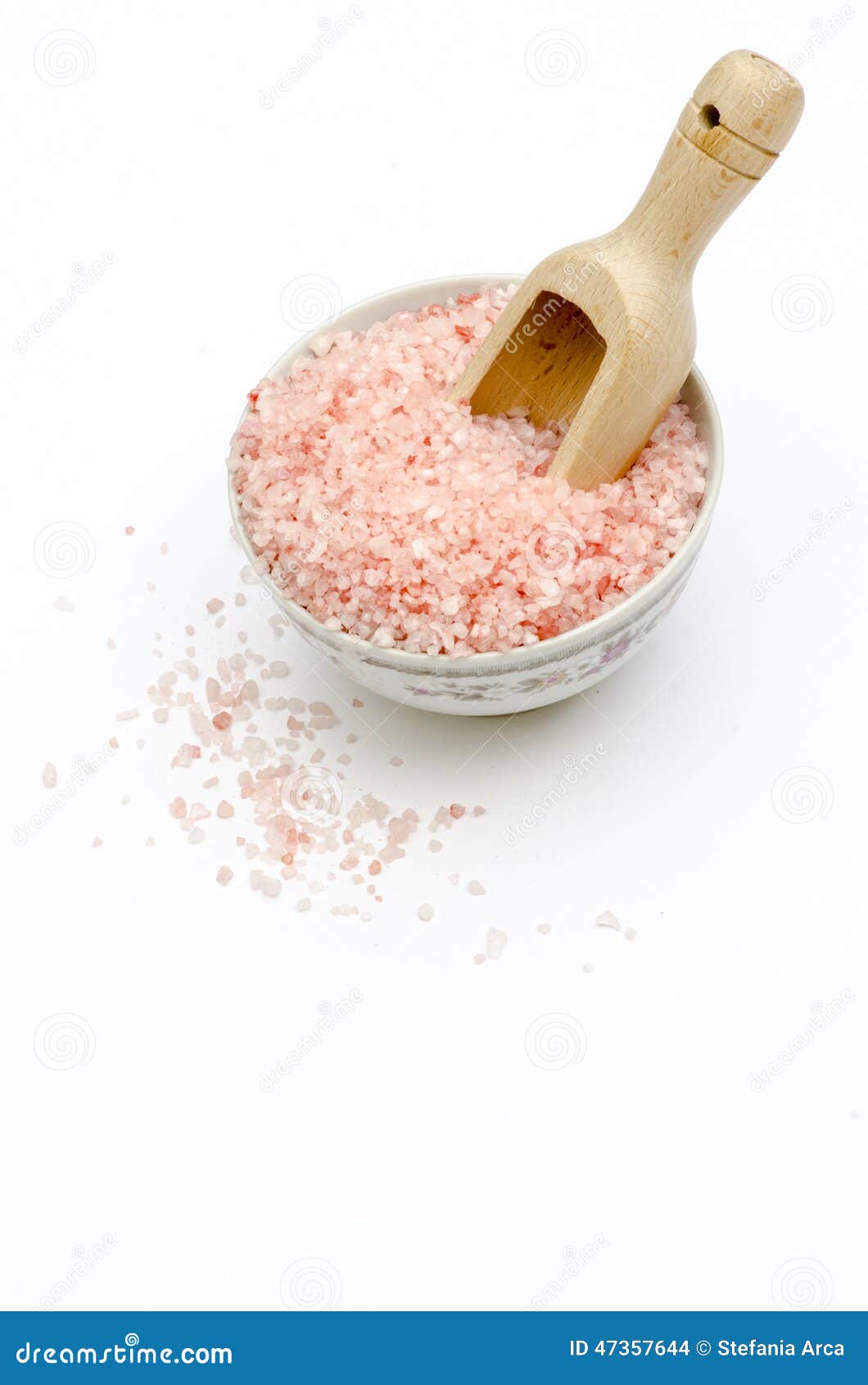 bowl full of pink bath salts, spoon, grains of salt fallen, on white background frontal view