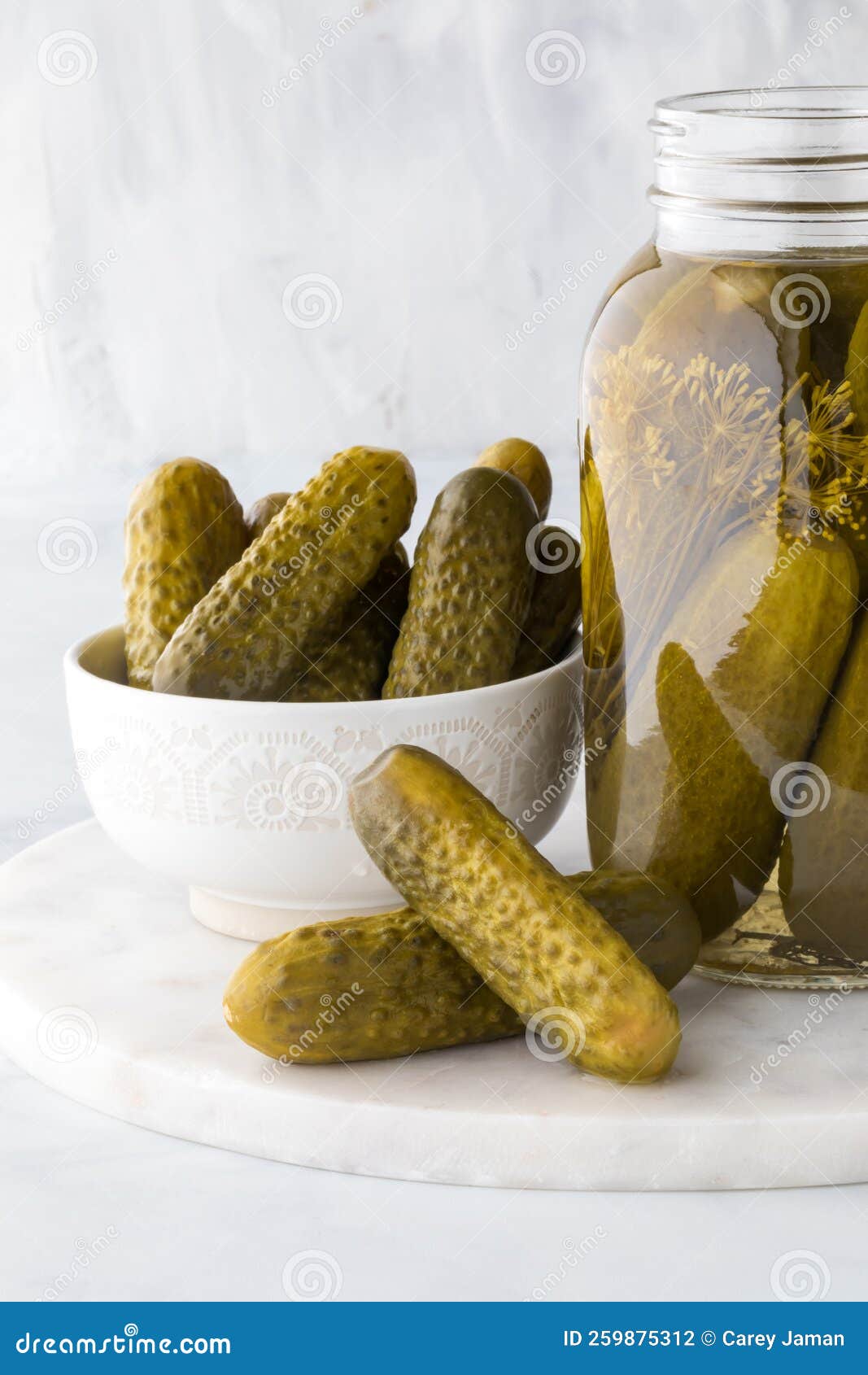 A Bowl and Jar of Dill Pickles with Two Pickles in Front on a Marble