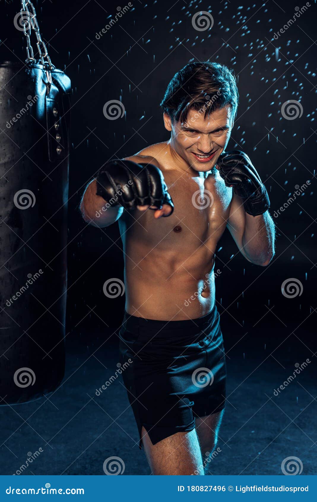 Boxeur De Sourire Dans Les Gants Debout Près Du Sac De Sable Sous Des  Gouttes D'eau Sur Le Noir. Photo stock - Image du entraînement, folâtre:  180827496