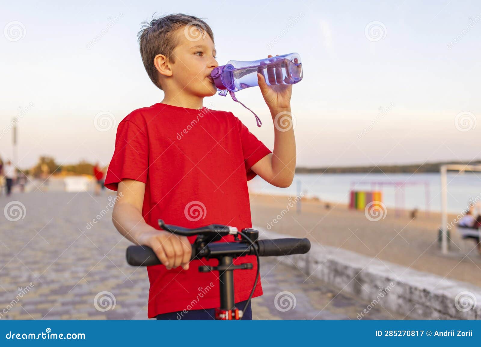 A Boy Drinks Water from Plastic Bottle, Standing with Scooter Stock