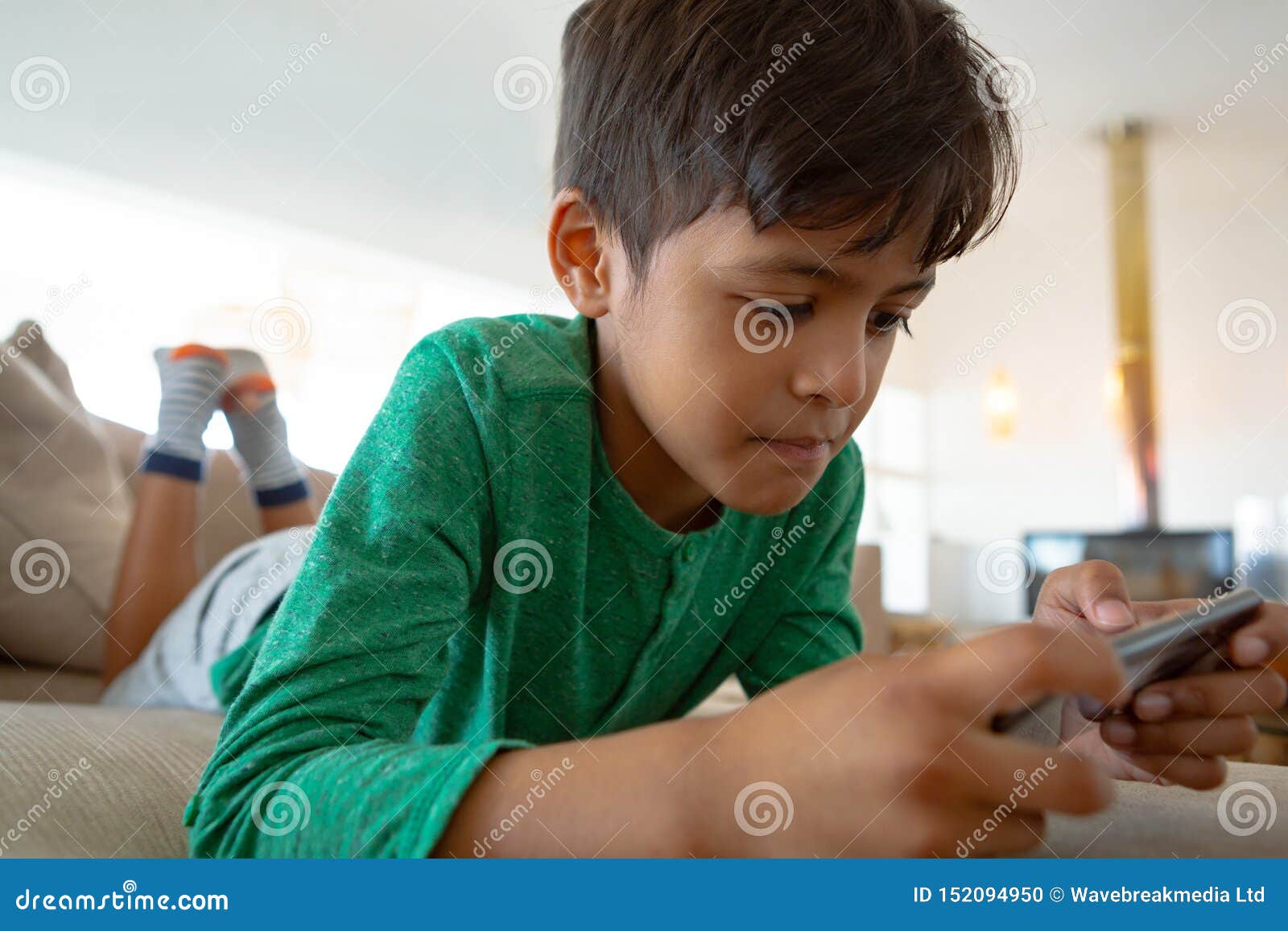 Boy Playing Game On Mobile Phone While Lying On Sofa At Home Stock