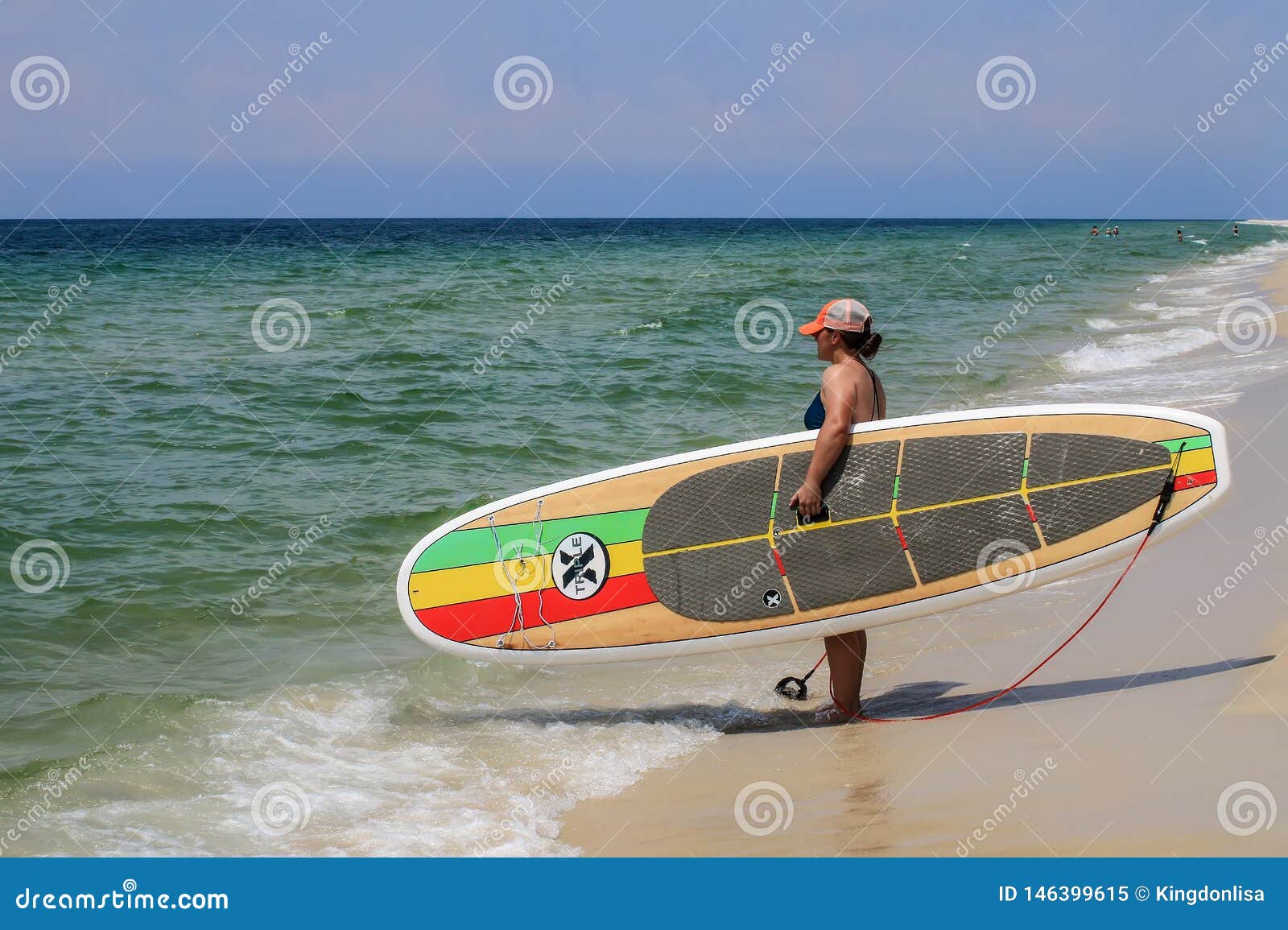 Bräde för skovel för surfareinnehavtrippel X på stranden. En ung kvinna som rymmer en trefaldig bambu för X ’Kontrol ’, står upp skovelbrädet på stranden med himlen och havet i bakgrunden
