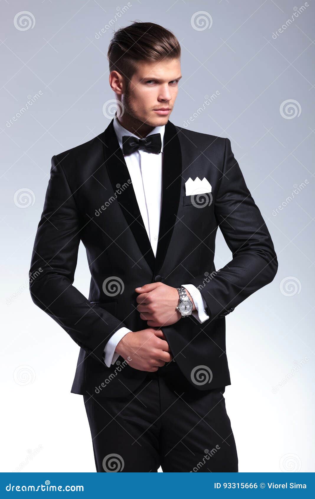 Business man adjusts his tux. Elegant young fashion man adjusting tuxedo with both hands while looking at the camera. on gray background