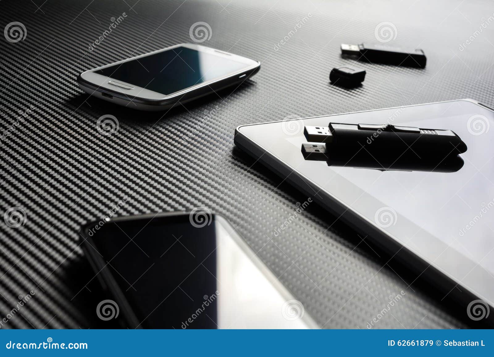 2 business mobiles with reflections and an usb drive lying next to a blank tablet with usb drive on top, all above a carbon layer