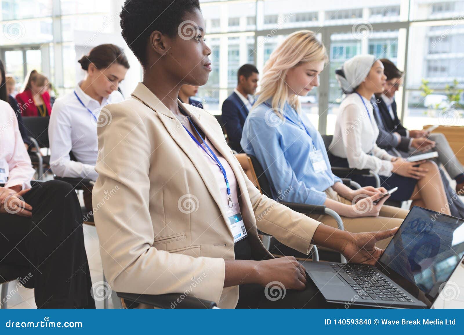  A group of people, including a woman in a beige suit using a laptop, are sitting in rows at an industry event.
