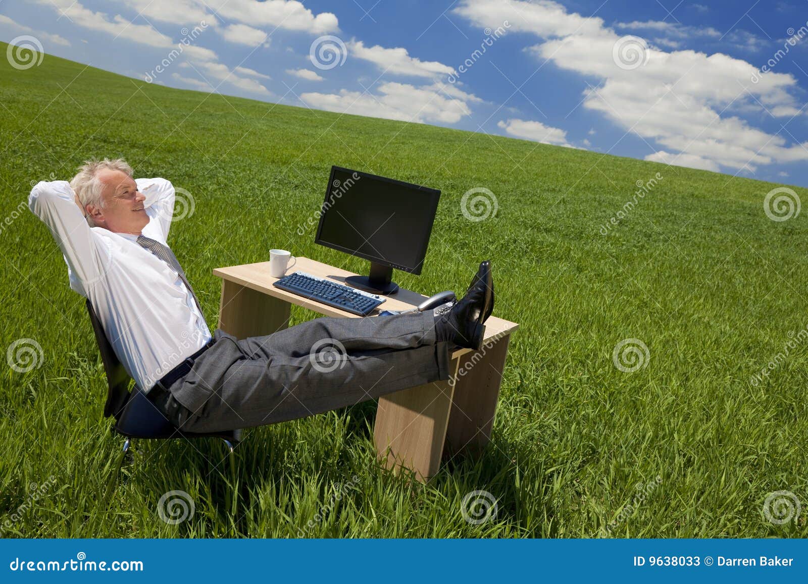 Businessman Relaxing In a Green Office. Business concept shot of a businessman relaxing with his feet up on his desk in a green field with a bright blue sky full of fluffy white clouds. Shot on location.