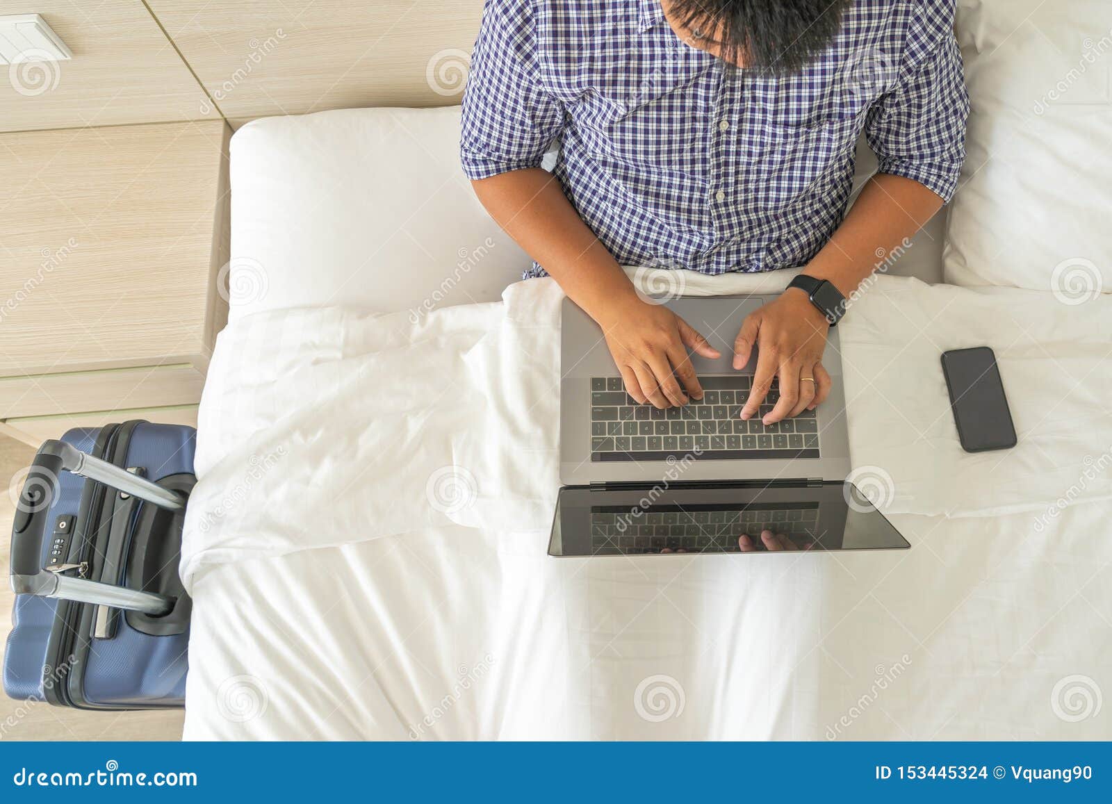 Businessman Using Laptop on Bed Next To Cellphone and Suitcase Stock
