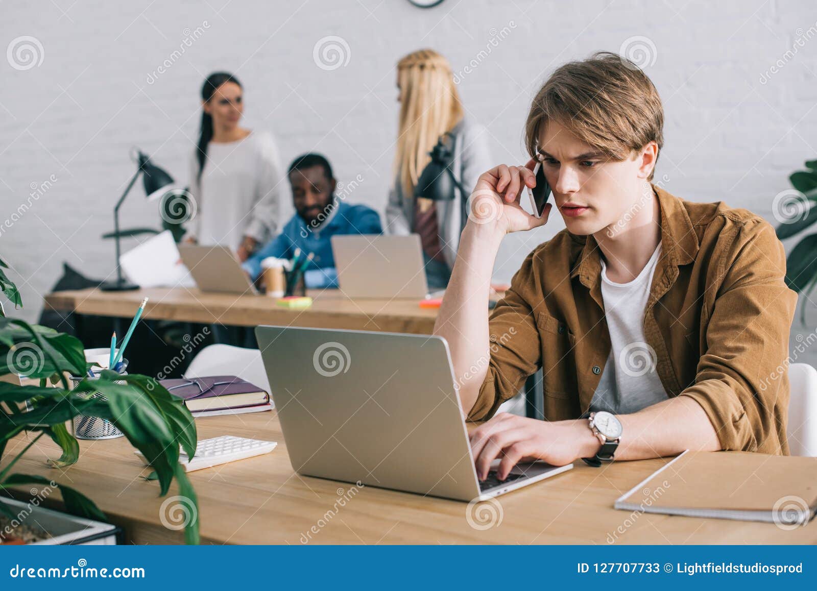 businessman using laptop and talking on smartphone and colleagues working behind