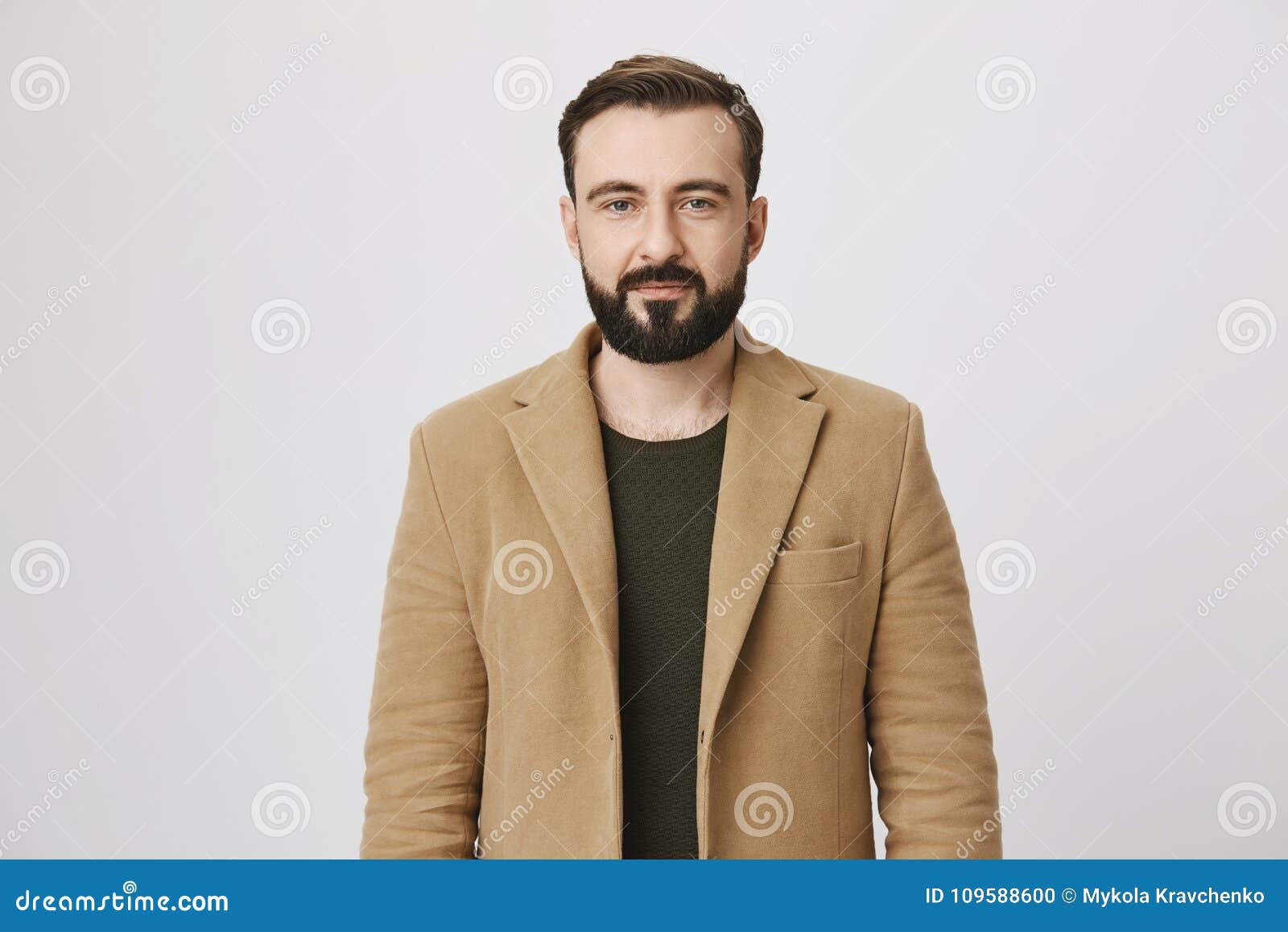 calm attractive bearded person in coat over green pullover with normal expression, standing against gray background. man