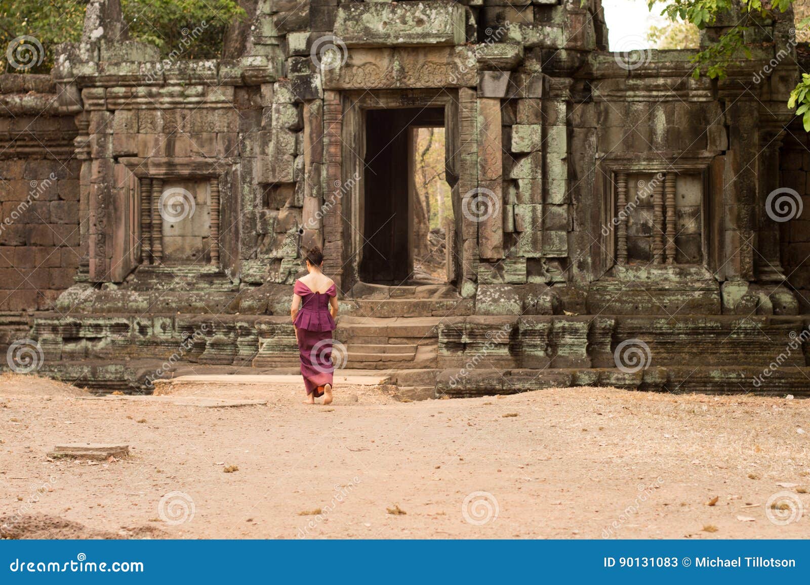 Cambodian Asian Girl in Traditional Dress Walks to the Doorway of a Temple Wall in Angkor. A Khmer girl in a traditional Khmer dress walks toward the doorway of an ancient wall at Angkor Thom.