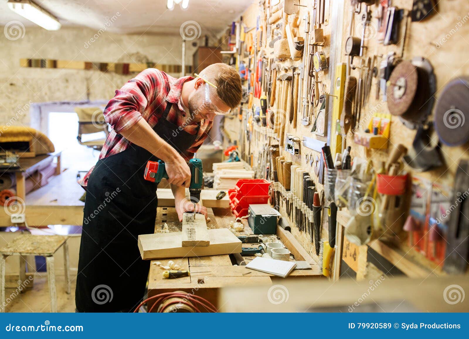 Carpentiere con la plancia di perforazione del trapano all'officina. Concetto di professione, della gente, di carpenteria, della lavorazione del legno e della gente - carpentiere con la plancia di legno di perforazione del trapano elettrico all'officina