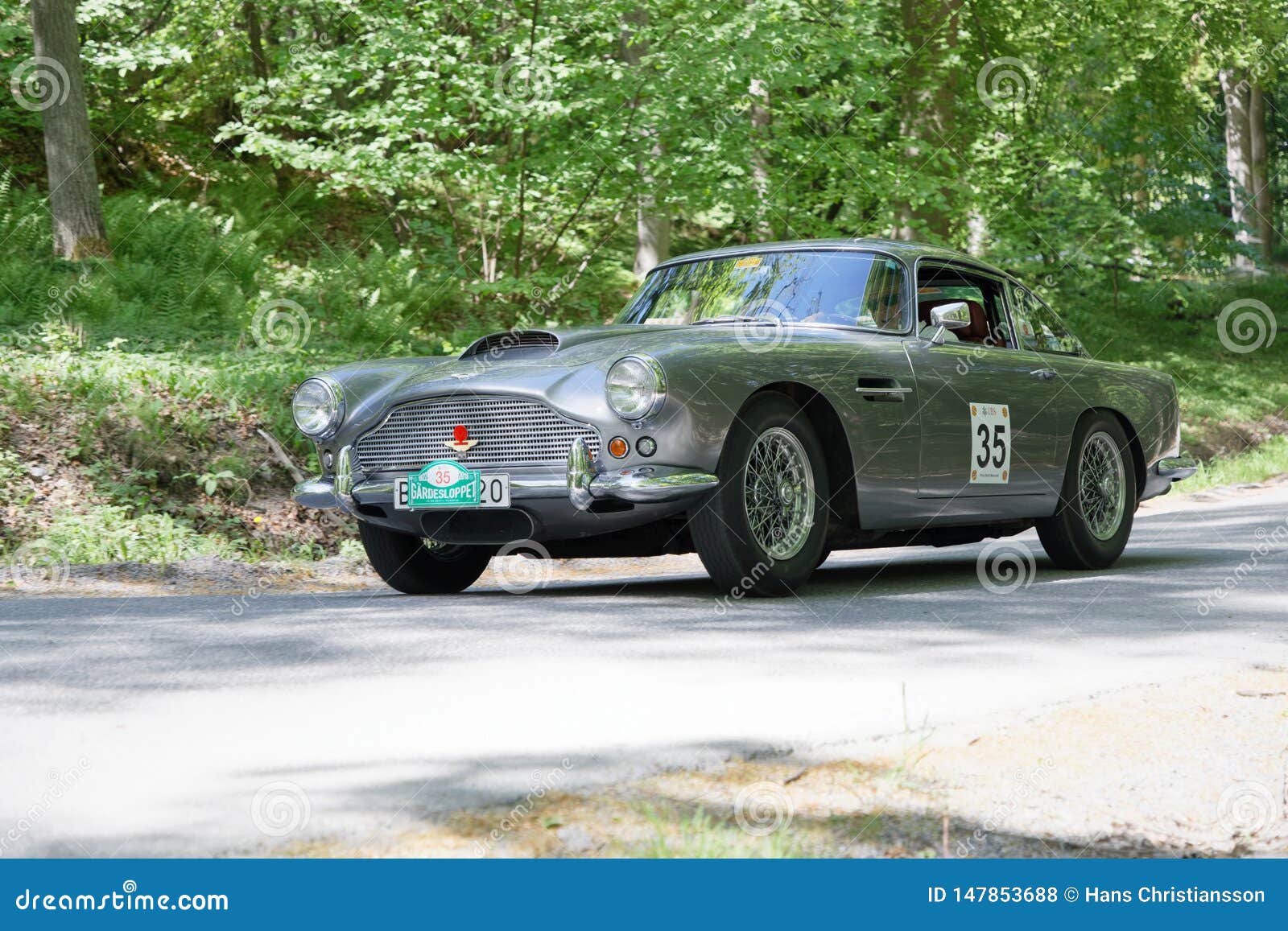 Carro clássico Aston Martin DB4 de Silver a cores de 1960 a conduzir numa estrada rural. ESTOCOLMO, SUÉCIA - 20 DE MAIO DE 2018: Carro clássico Aston Martin DB4 de Silver a partir de 1960, dirigindo na corrida pública Gardesloppet nas florestas de Djurgarden, Estocolmo, Suécia 20 de maio de 2018