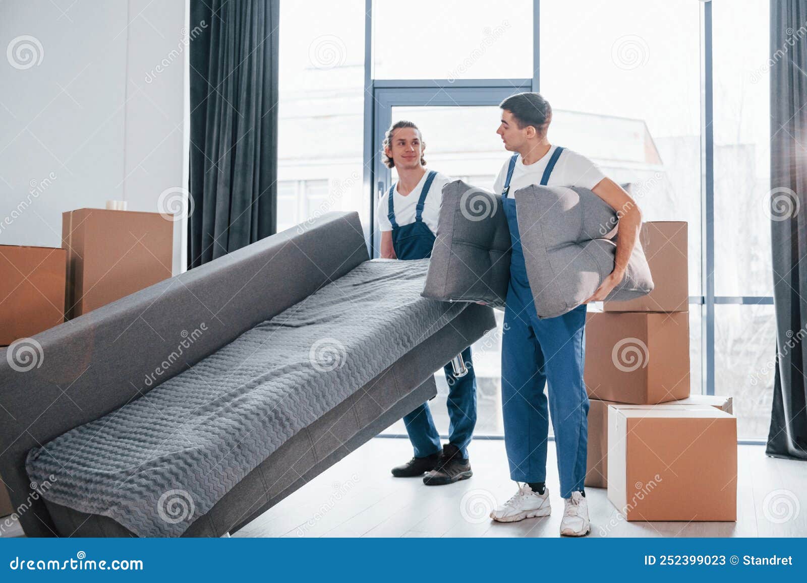 Carrying Heavy Sofa. Two Young Movers in Blue Uniform Working Indoors