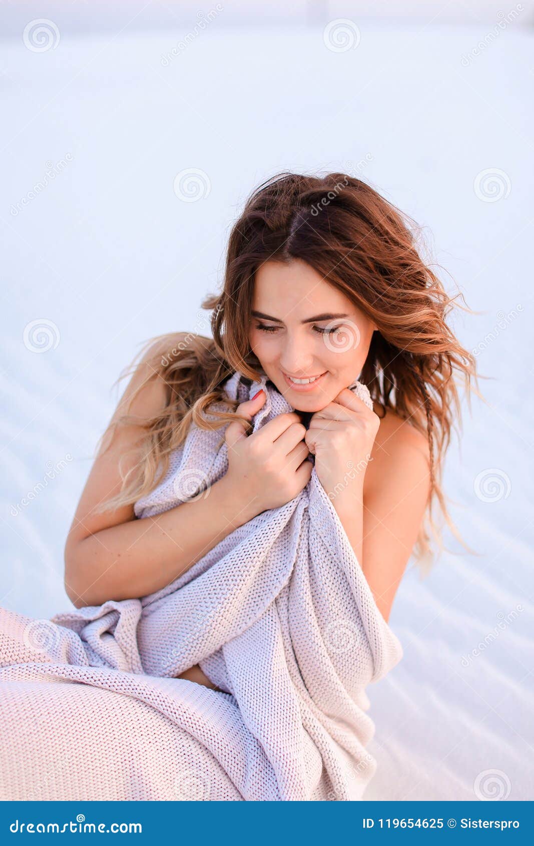 Caucasian female person wrapped in blanket sitting on white sand on beach. Concept of photo session in desert and relax on nature.
