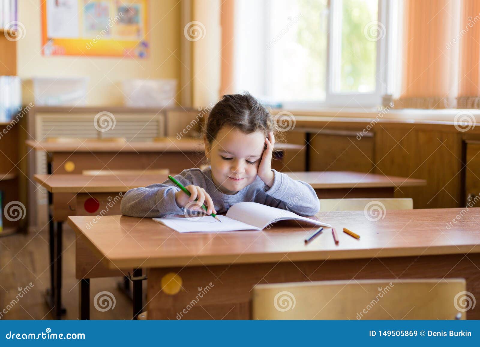 Caucasian Little Girl Sitting At Desk In Class Room And Begins To