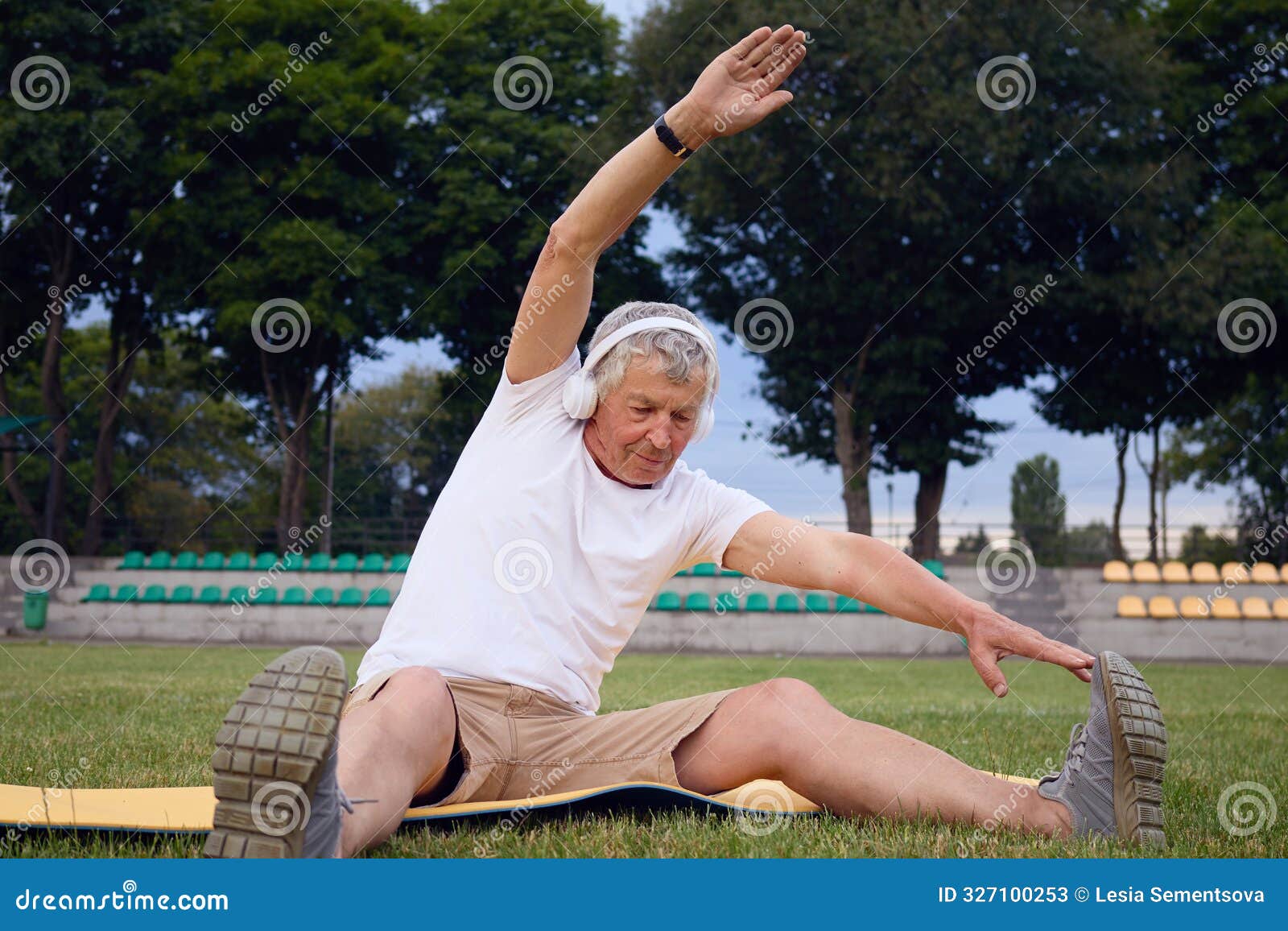 caucasian senior athlete in a white t-shirt and wireless headphones sits on yoga mat at stadium stretching his legs and relishing