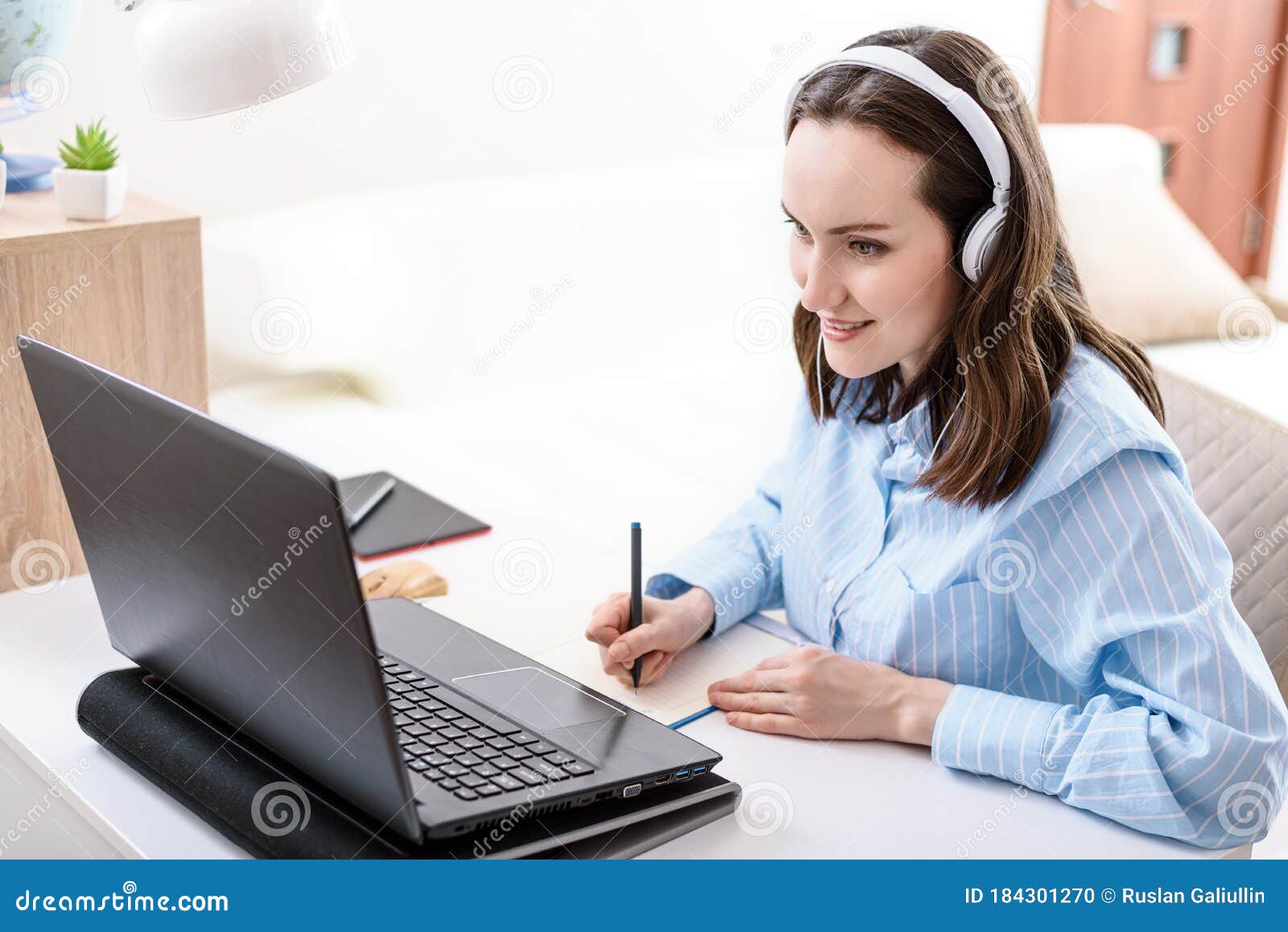 caucasian woman with headphones writes in notebook, looks at laptop screen, remote work at home, home training