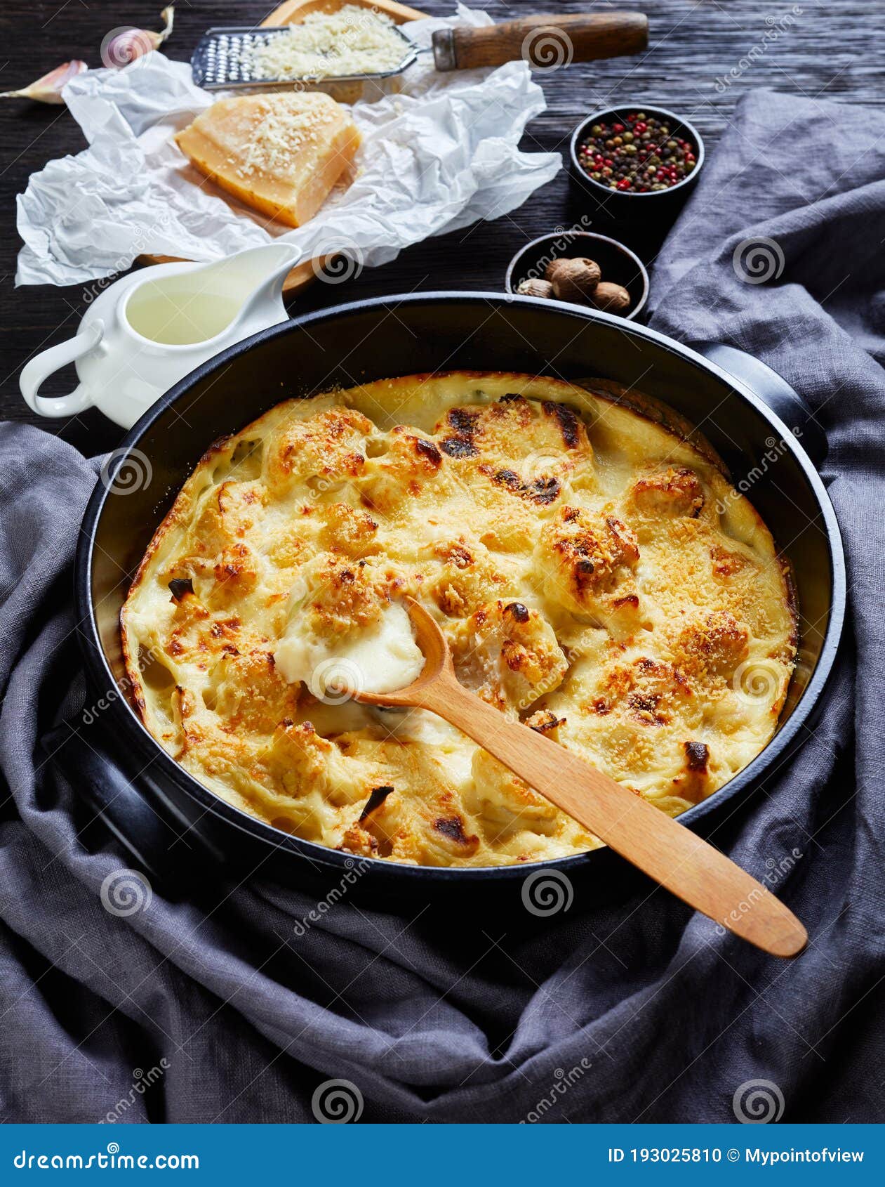 Cauliflower cheese casserole on a black dish. Cauliflower cheese casserole- vegetable side dish served on a black baking dish on wooden background with ingredients: garlic, nutmeg, peppercorns, milk, parmesan cheese, close-up