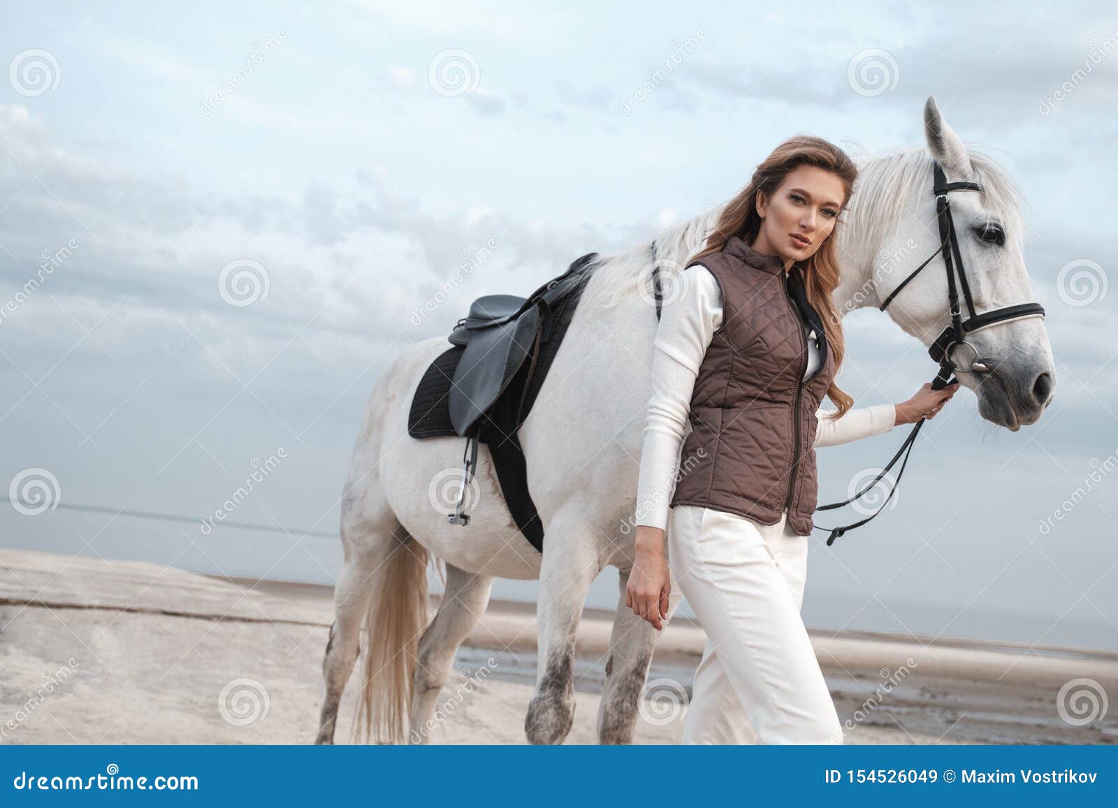 charming and beautiful young woman wearing stylish jockey outfit is holding the reins and posing with the white horse