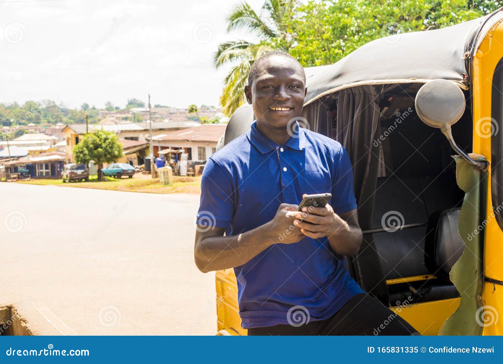 cheerful african man standing next to his tuk tuk taxi smiling and using his smart phone