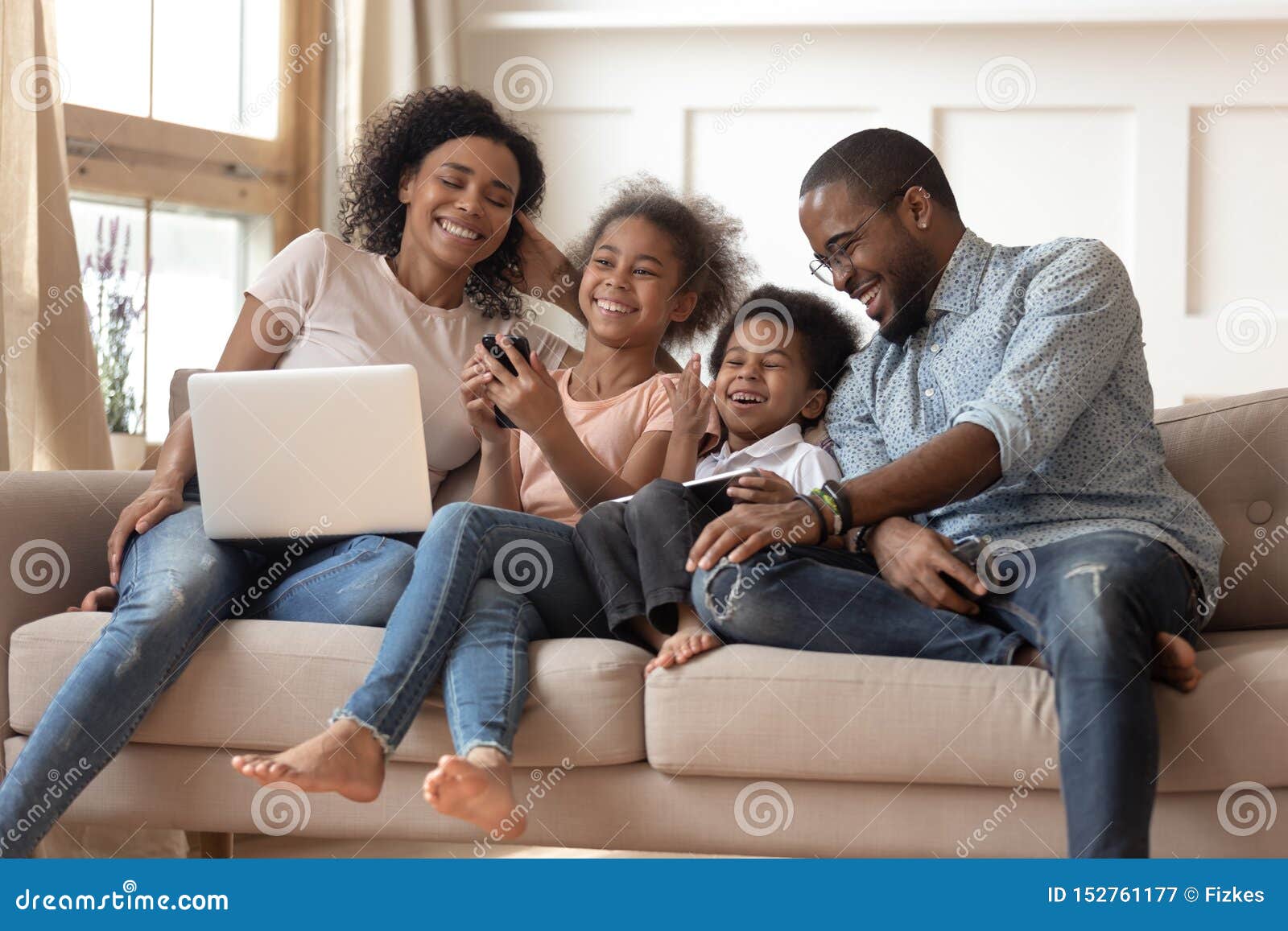 cheerful african parents and kids laugh using devices on sofa