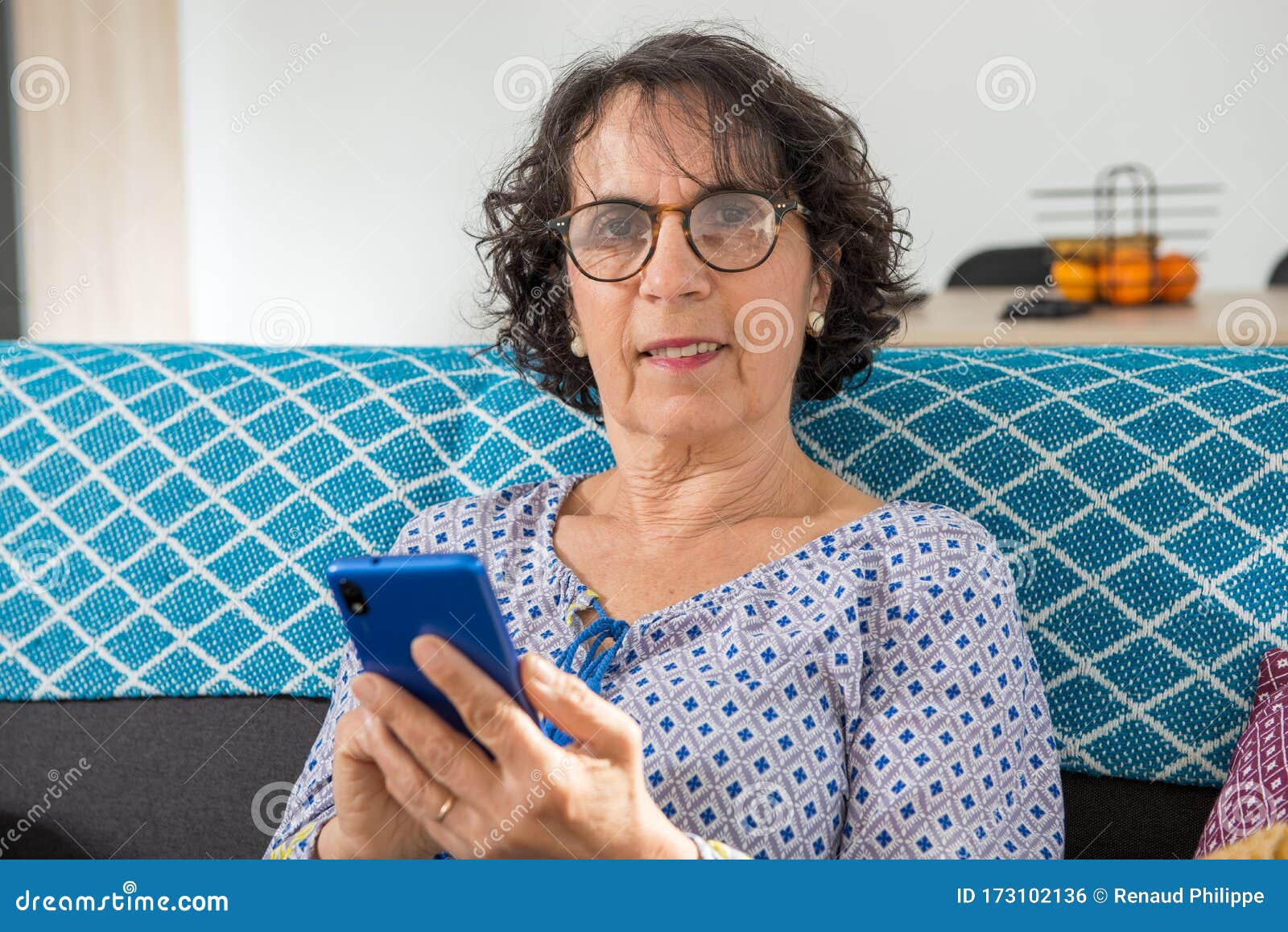 cheerful brunette senior woman using smartphone while sitting on sofa