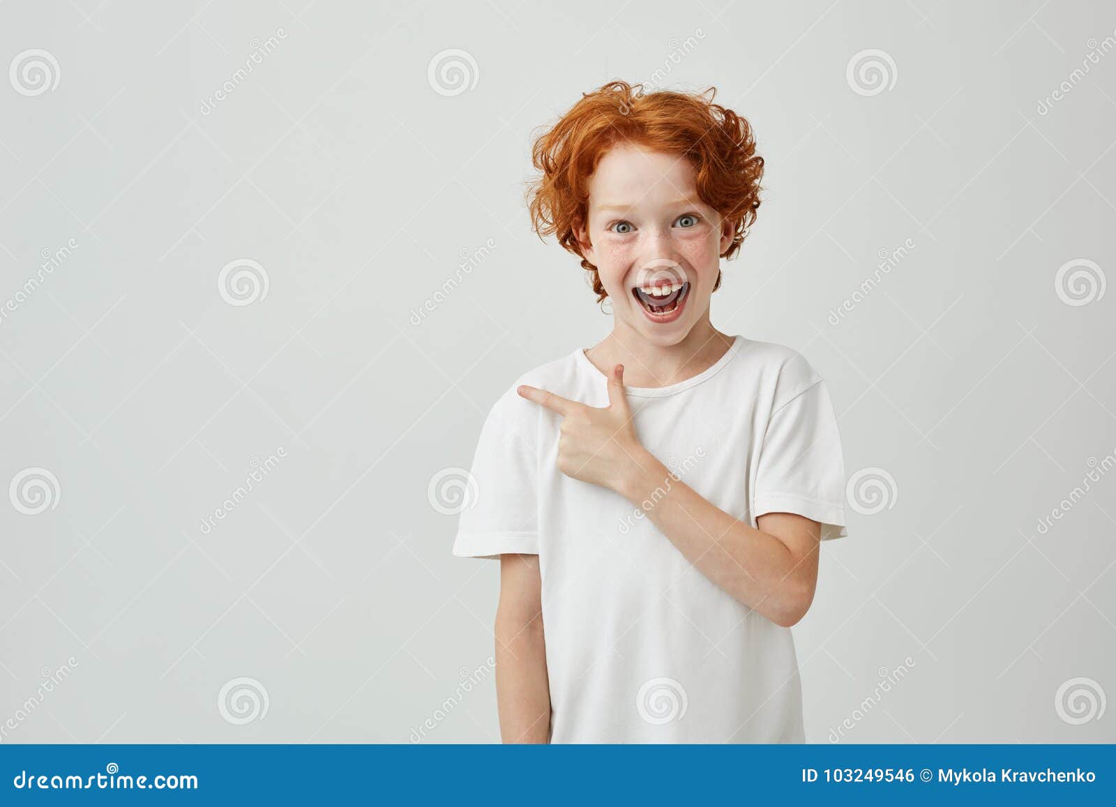 cheerful cute little boy with curly ginger hair and freckles happy smiling and pointing aside with finger on white