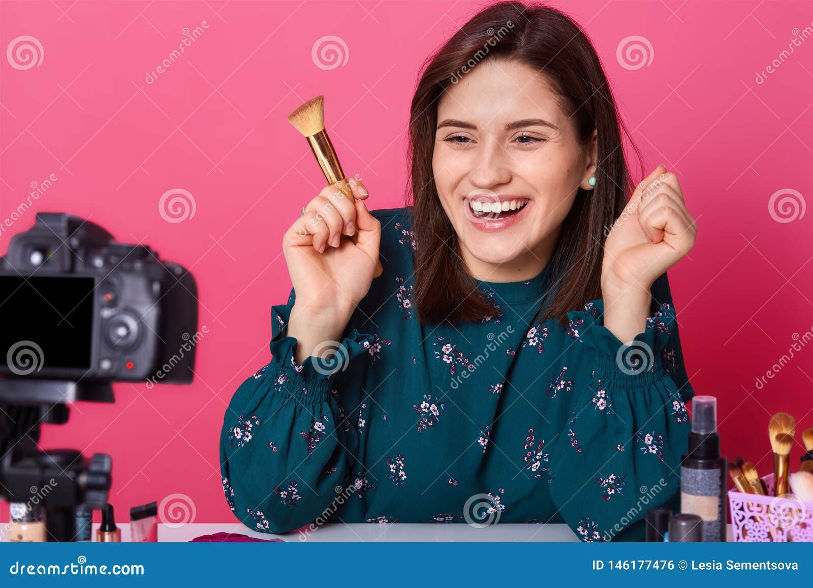 cheerful magnetic brunette blogger posing  over pink background, clenches her fists, holding brush for makeup in one hand