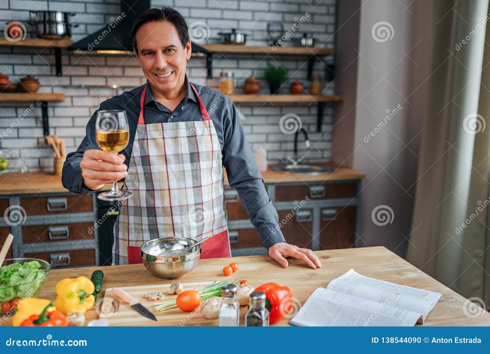 Cheerful Man Stand at Table in Kitchen. he Hold Glass of White Wine and ...