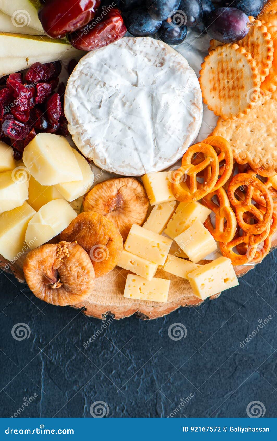 cheese plate grape salted craker cookies dry dates cranberry, fig, pear pretzels on a wooden board on a black slate background.