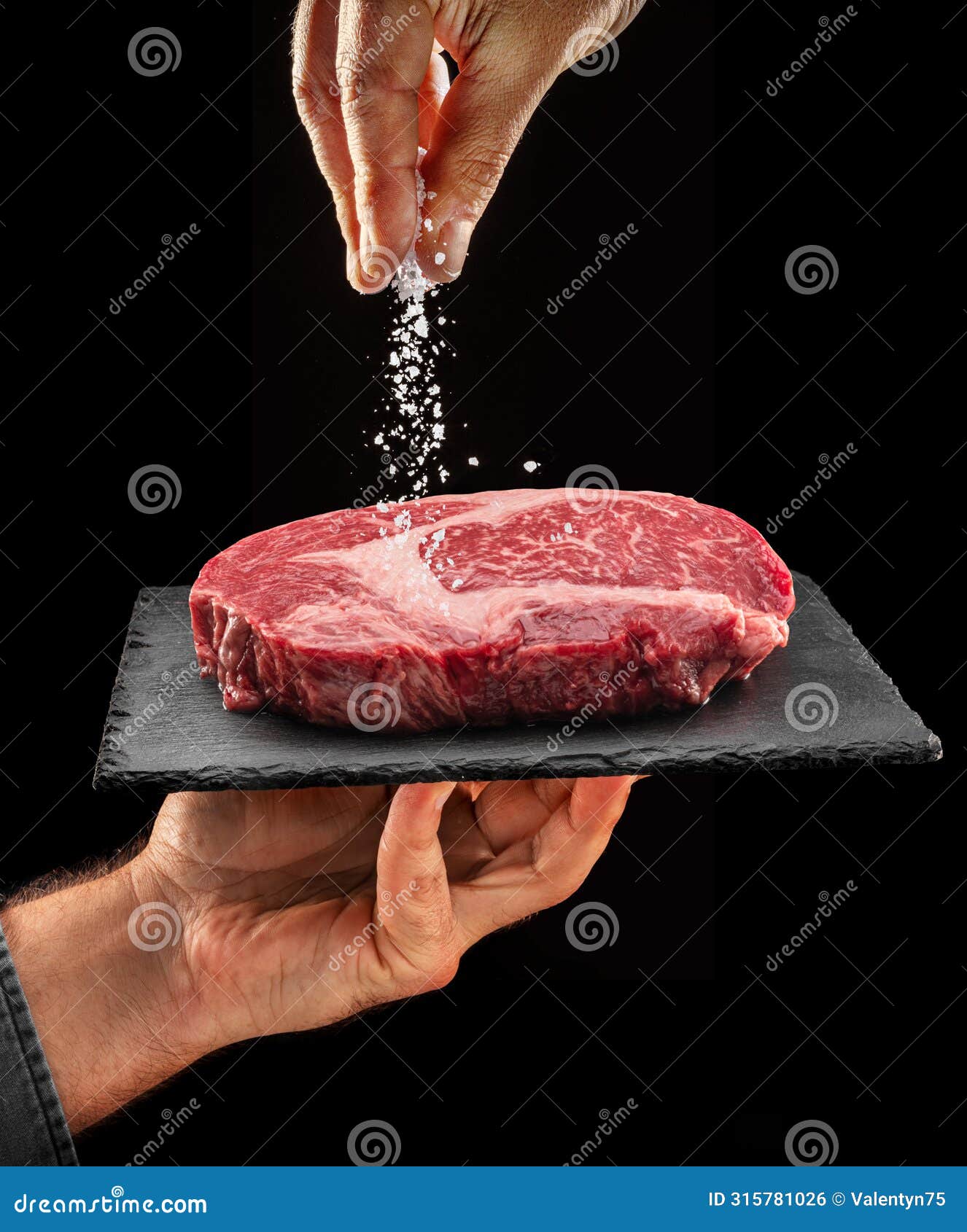 chef is salting or seasoning raw ribeye steak laying on graphite serving board. black background