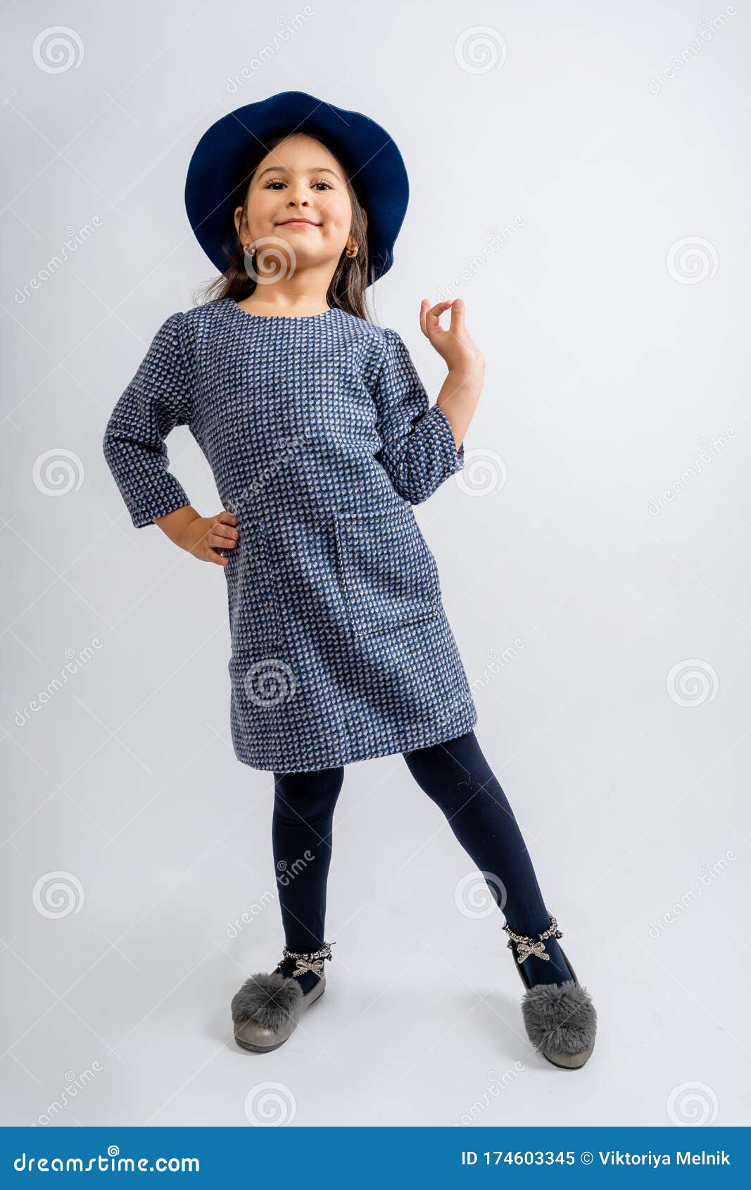 a child in a blue dress with white and black spots, a blue hat, blue tights and gray shoes  on a white background.