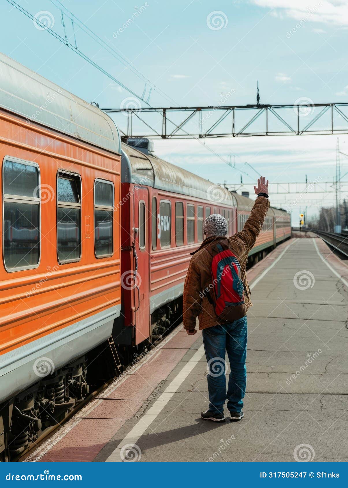 a child bundled up in winter clothing extends a hand high to wave at a departing orange train on a clear day.