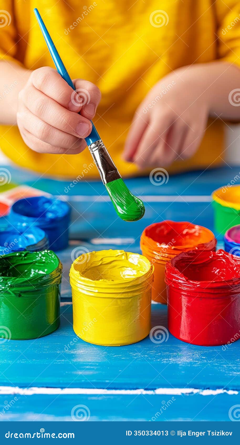 child s hand dips paintbrush into vibrant yellow pot, surrounded by colorful paints, fun art concept