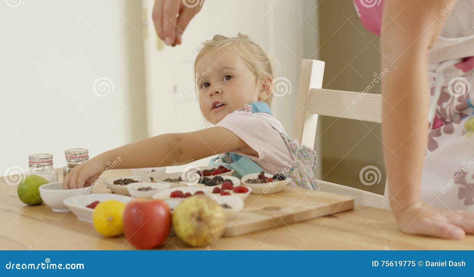 Child watching woman sprinkle candy on muffins. Cute blond female child watching woman leaning on table while sprinkling candy sprinkles on muffins to be baked
