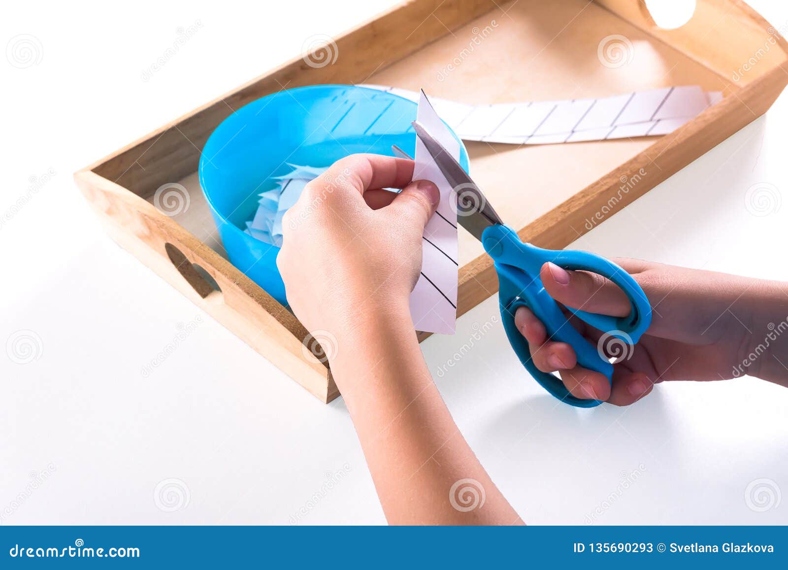 children`s hands hold blue scissors and cut the paper. on a wooden tray are montessori materials for a lesson