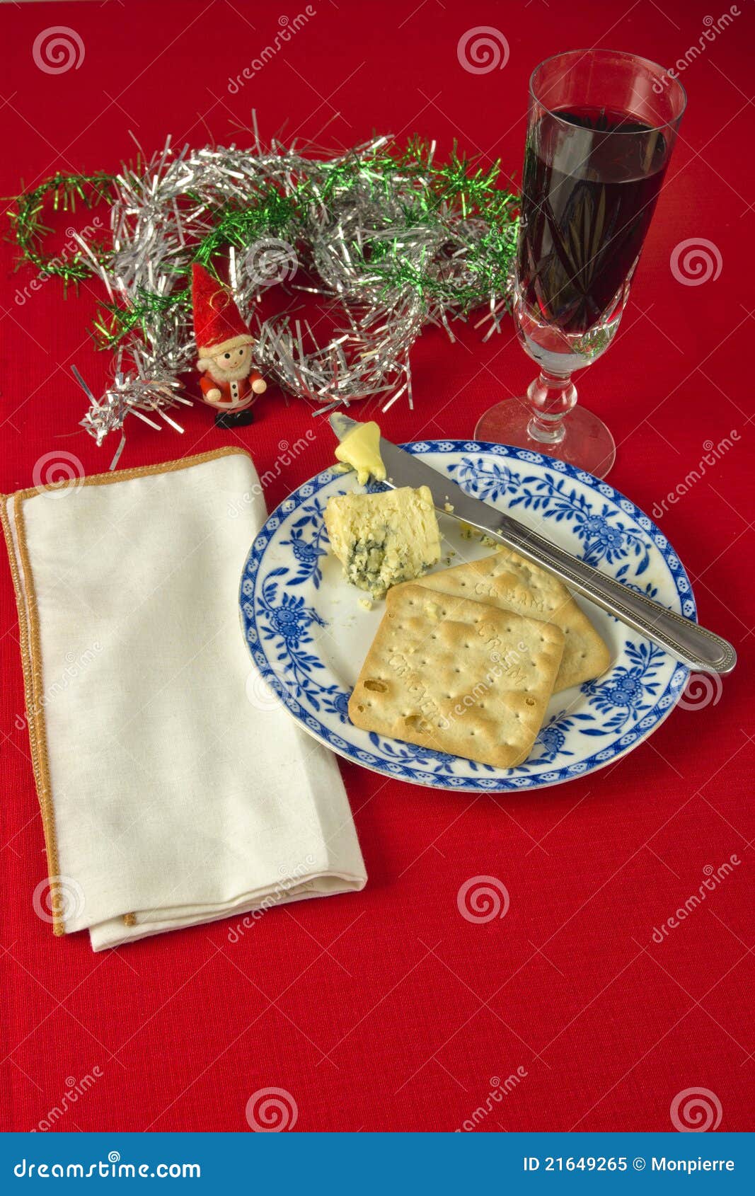 Christmas Cheese and Wine. Two crackers and Stilton cheese on blue and white plate , with knife and butter. White napkin against red table cloth, with decorations.Glass of red wine.