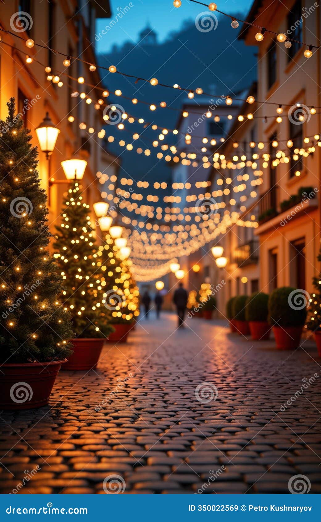 christmas decorated city street at dusk. small christmas trees with warm lights lining cobblestone street. festive string lights