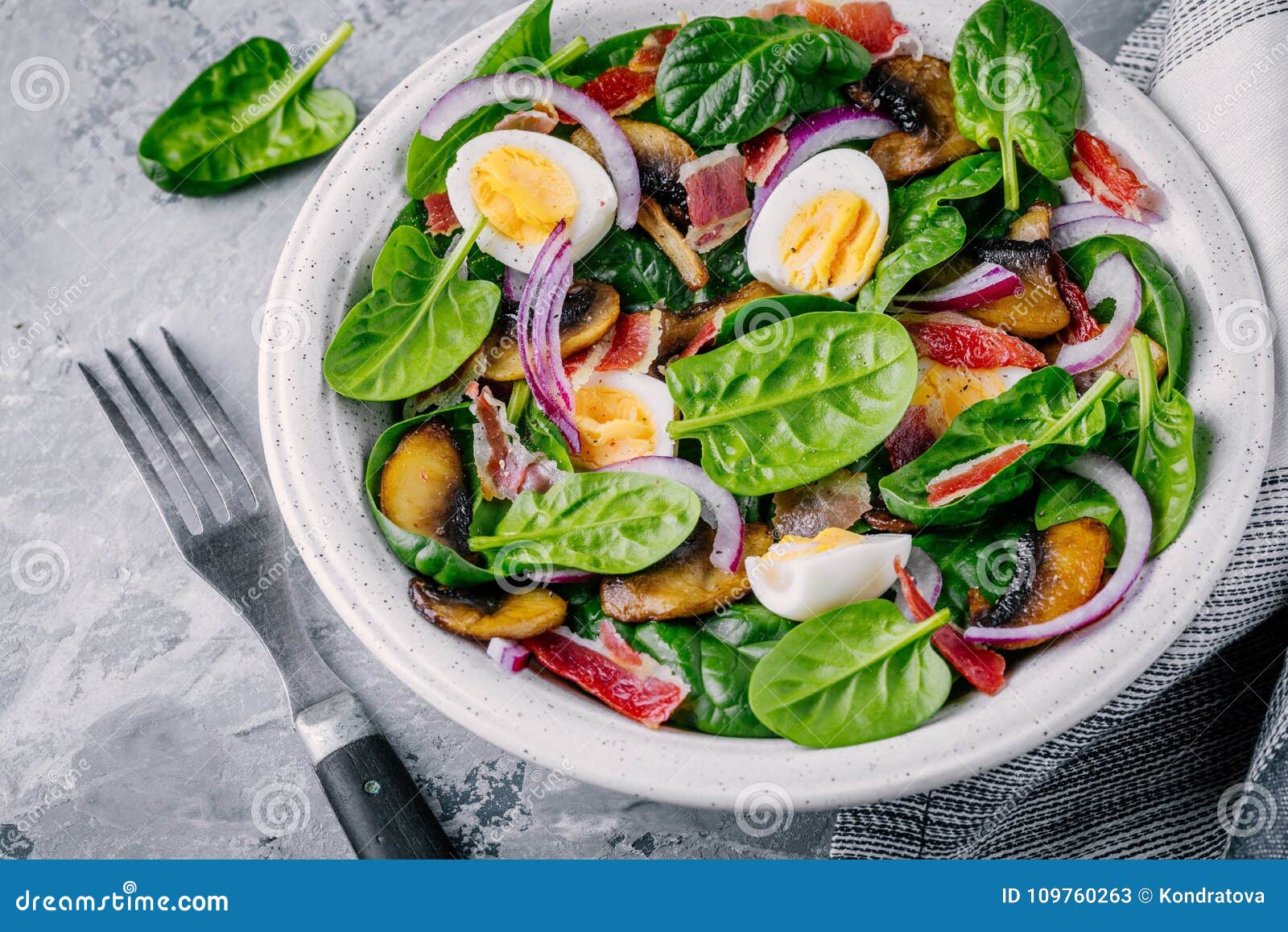 Ciotola del pranzo di insalata degli spinaci con bacon, i funghi, le uova e le cipolle rosse su fondo grigio