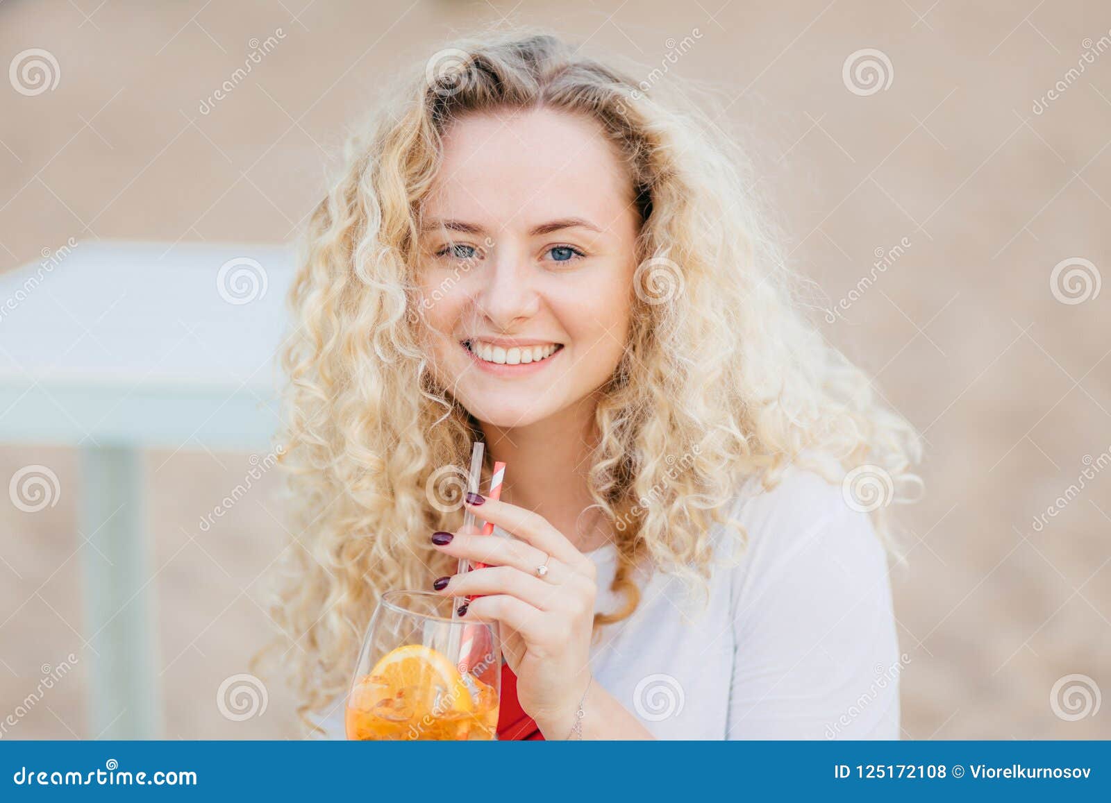 Ciérrese encima del retrato de la hembra joven atractiva con la expresión positiva, dientes perfectos blancos de las demostraciones, cóctel fresco de las bebidas, actitudes contra fondo de la playa, se divierte con los amigos, disfruta de tiempo de verano