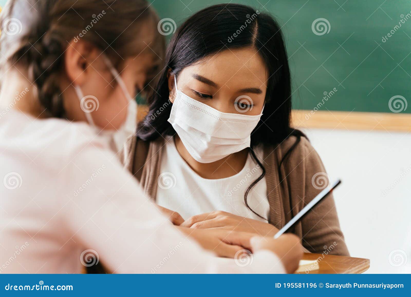 close-up of asian female teacher wearing a face mask in school building tutoring a primary student girl. ary