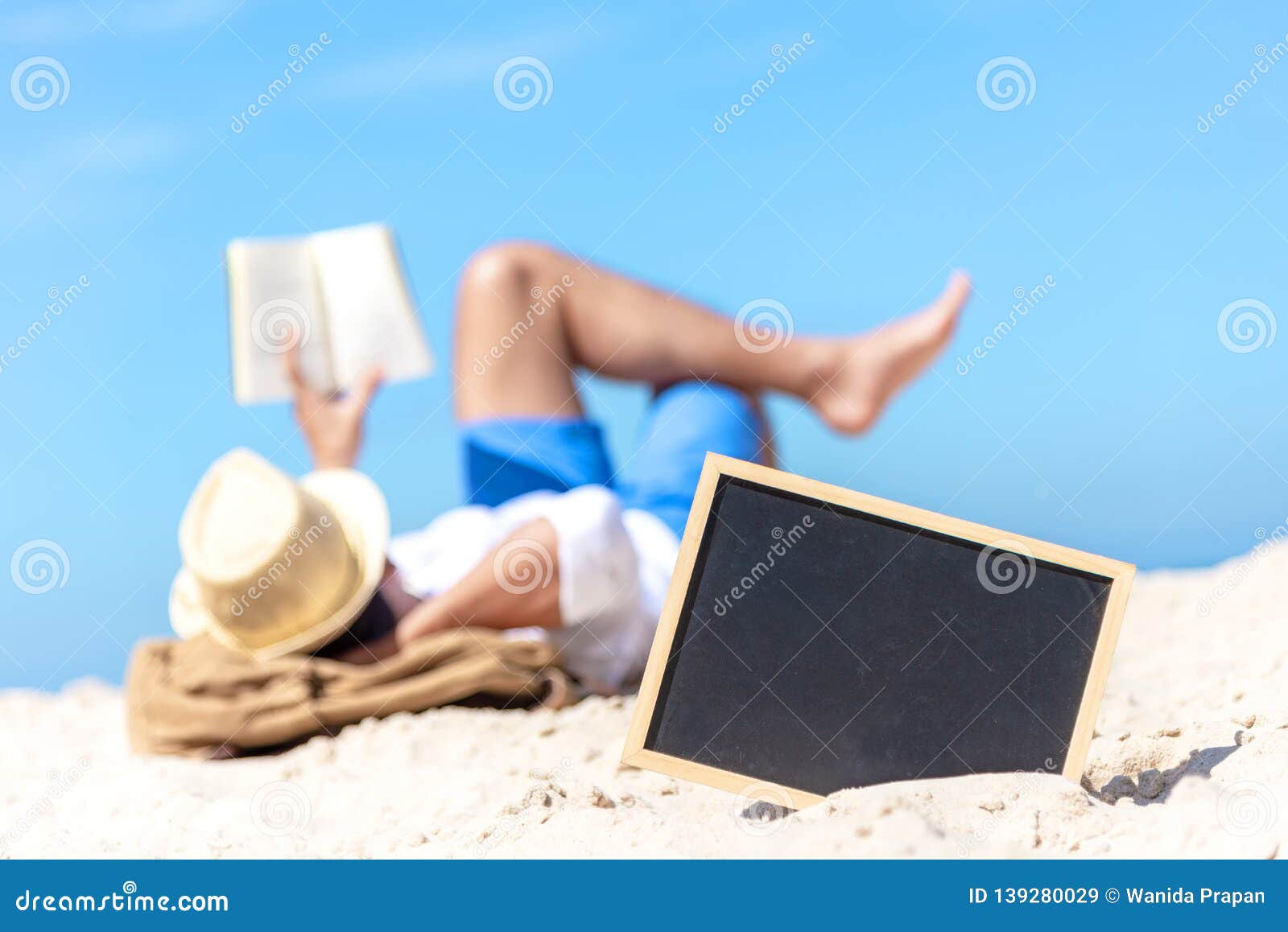 close up of a chalkboard on the sand of a beach, background happy smiling caucasian tourist asian young man relax and reading book