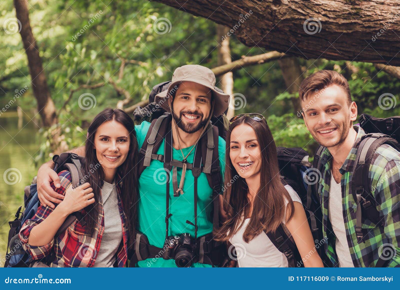 Close up cropped portrait of four cheerful friends in the summer nice wood. They are hikers, walking and picking place for camping, embracing, posing for photo